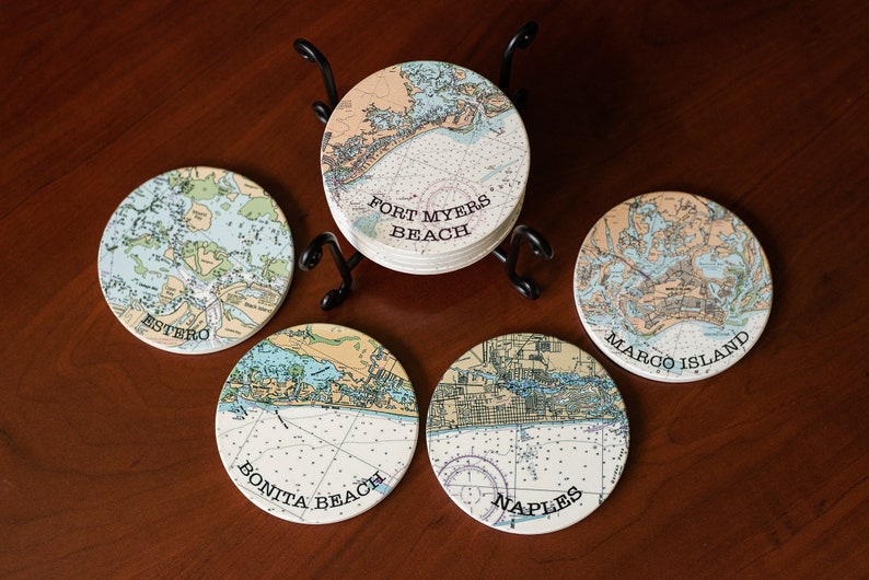 May include: Set of six round ceramic coasters featuring nautical maps of different Florida locations. The coasters are arranged in a circle on a wooden surface with a metal stand in the center. The coasters feature the names "Estero", "Bonita Beach", "Naples", "Marco Island", and "Fort Myers Beach".