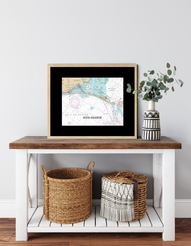 May include: A framed nautical chart of Boca Grande, Florida, with a black frame and a white mat. The chart shows the coastline, waterways, and surrounding islands. The chart is displayed on a wooden console table with two woven baskets.