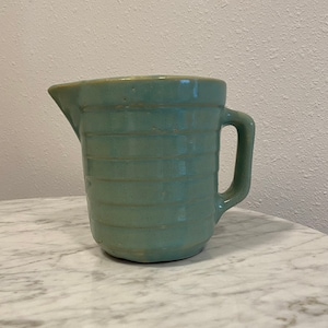 May include: A light green ceramic pitcher with a handle. The pitcher has a ribbed design and a slightly rounded base.