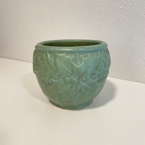 May include: A light green ceramic planter with embossed floral and berry designs. The round planter has a slightly flared rim and a textured surface. The color is a muted, pastel green.