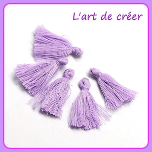 May include: Six lavender-colored fabric tassels, each with a knotted top and a fringe of soft fibers.  "L'art de créer" is written in black text at the top of the image.