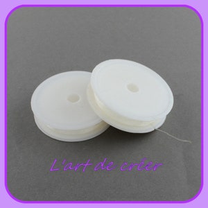 Peut inclure: Deux bobines de fil élastique blanc, chacune enroulée autour d'une bobine en plastique blanc avec un trou au centre. Les bobines sont posées sur une surface grise. L'art de créer est écrit en texte violet sous les bobines.