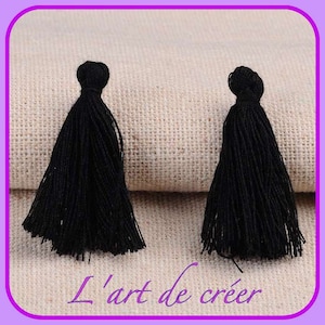 Peut inclure: Deux glands noirs, chacun avec une petite boucle en haut, sont présentés sur un fond en tissu beige. Les glands mesurent environ 5 cm de long et ont une texture douce et moelleuse.