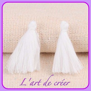 Peut inclure: Deux pompons blancs et moelleux sur un fond de tissu beige. Les pompons mesurent environ 5 cm de long et ont une frange en bas.