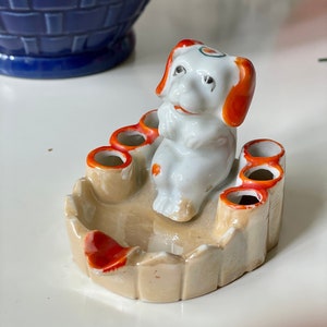 以下が含まれることがあります： オレンジ色のアクセントが付いたベージュの灰皿に座っている白い磁器製の犬の置物。灰皿には、縁の周りにタバコ用の小さな穴が6つあります。