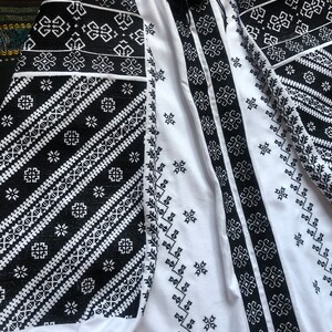 May include: White blouse with black embroidered patterns. The long sleeves and front panels feature intricate geometric designs. A black tassel hangs from the neckline. The blouse is made of a lightweight fabric.