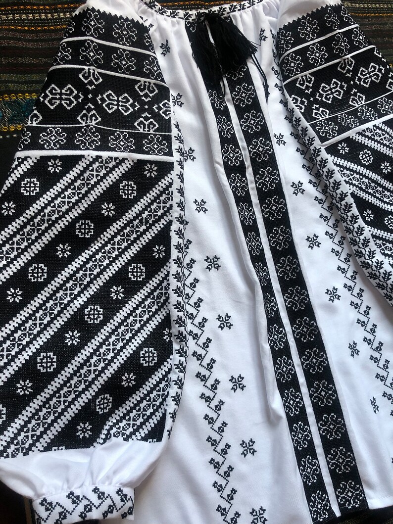 May include: White blouse with black embroidered patterns. The long sleeves and front panels feature intricate geometric designs. A black tassel hangs from the neckline. The blouse is made of a lightweight fabric.