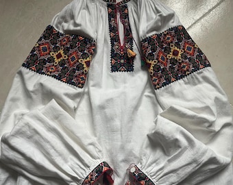 May include: White long-sleeved blouse with intricate embroidery in shades of red, orange, yellow, and green on the sleeves and neckline. The blouse features a loose fit and tassel details, showcasing a traditional folk design.