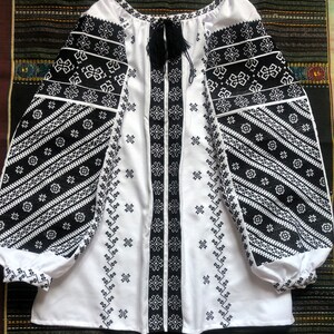 May include: White blouse with black embroidered details. The long sleeves and front panel feature intricate geometric patterns. A black tassel tie adorns the neckline. The blouse is made of a soft, flowing fabric.