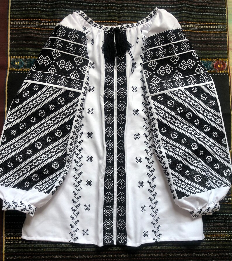 May include: White blouse with black embroidered details. The long sleeves and front panel feature intricate geometric patterns. A black tassel tie adorns the neckline. The blouse is made of a soft, flowing fabric.