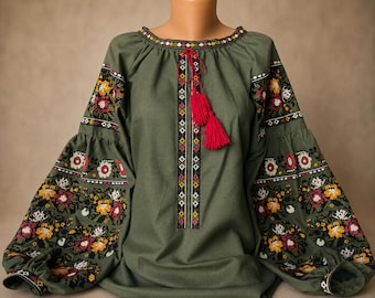 Puede incluir: Una blusa de manga larga color verde oliva con intrincados bordados florales en tonos amarillo, rojo y blanco en las mangas y el cuello. La prenda incluye un panel central vertical bordado y un cordón rojo con dos borlas.