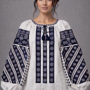 May include: White blouse with navy blue floral embroidery on the sleeves and bodice. The long sleeves have a puffy design at the wrist. The neckline and bodice feature intricate navy blue patterns and a tassel tie.