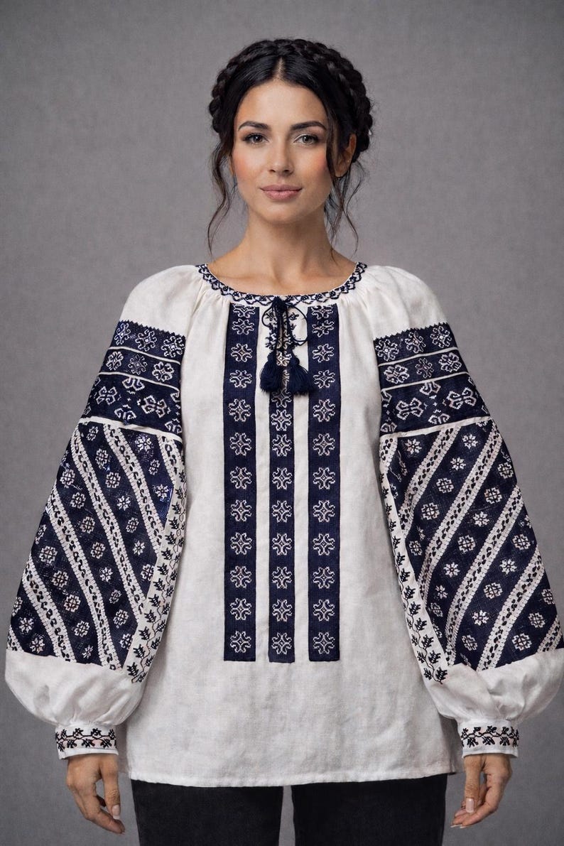 May include: White blouse with navy blue floral embroidery on the sleeves and bodice. The long sleeves have a puffy design at the wrist. The neckline and bodice feature intricate navy blue patterns and a tassel tie.