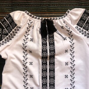 May include: White embroidered blouse with black geometric patterns on the sleeves, neckline, and front. The blouse features a drawstring neckline with black tassels. The embroidery is detailed and the fabric appears to be cotton.