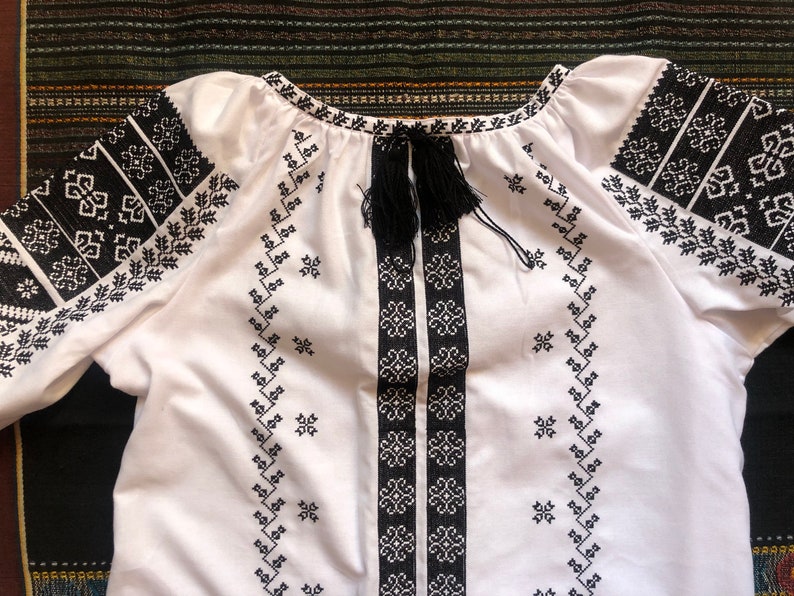 May include: White embroidered blouse with black geometric patterns on the sleeves, neckline, and front. The blouse features a drawstring neckline with black tassels. The embroidery is detailed and the fabric appears to be cotton.