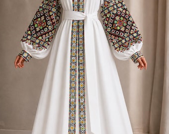 May include: A floor-length white dress featuring long, voluminous sleeves and a V-neckline. The garment is adorned with intricate, multi-colored geometric embroidery along the front placket and sleeves, and includes a matching white fabric waist tie.