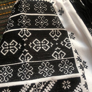 May include: White blouse with black embroidered floral patterns on the sleeves and neckline. The embroidery features geometric and floral designs. The sleeves have black fringe. The blouse is made of a lightweight fabric.