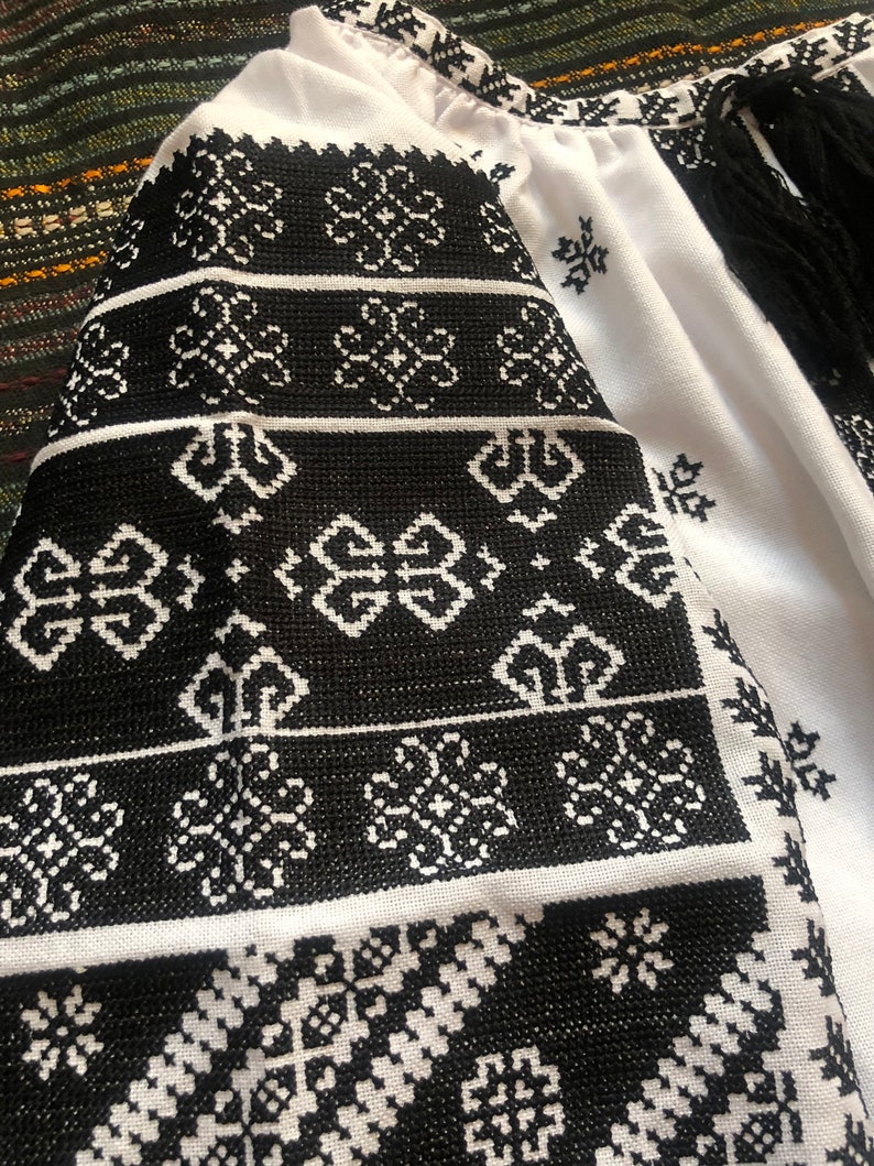May include: White blouse with black embroidered floral patterns on the sleeves and neckline. The embroidery features geometric and floral designs. The sleeves have black fringe. The blouse is made of a lightweight fabric.