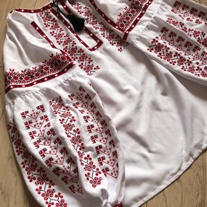 Könnte beinhalten: Weiße bestickte Bluse mit roten und schwarzen Blumenmustern. Die Bluse hat lange Ärmel mit einem gerafften Bündchen und einem runden Ausschnitt.