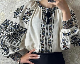 May include: White linen blouse with black, blue, and red embroidered floral patterns. The blouse has long sleeves with embroidered cuffs and a round neckline.