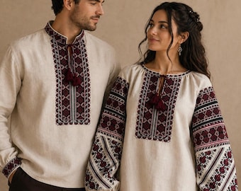 May include: Beige linen shirts with intricate burgundy and black embroidery. The shirts feature long sleeves and a detailed pattern around the neckline and cuffs. The woman's shirt has a tassel tie.
