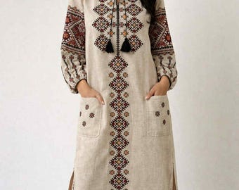 May include: A beige linen maxi dress with intricate embroidered details in shades of red, brown, and gold. The dress features long sleeves, pockets, and a split hem. The neckline is adorned with tassels.