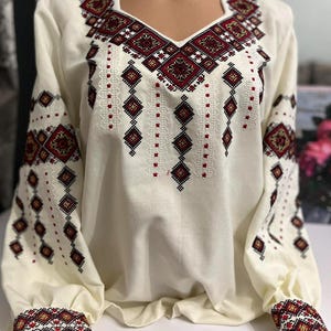May include: Cream-colored long-sleeved blouse with intricate geometric embroidery in red, black, and gold. The embroidery adorns the neckline, sleeves, and cuffs, creating a detailed and traditional design. The blouse has a V-neck and a relaxed fit.