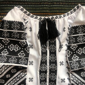 May include: White blouse with black embroidered floral patterns on the sleeves and bodice. The blouse features a round neckline and a black tassel tie. The sleeves are long and the fabric appears to be cotton or linen.