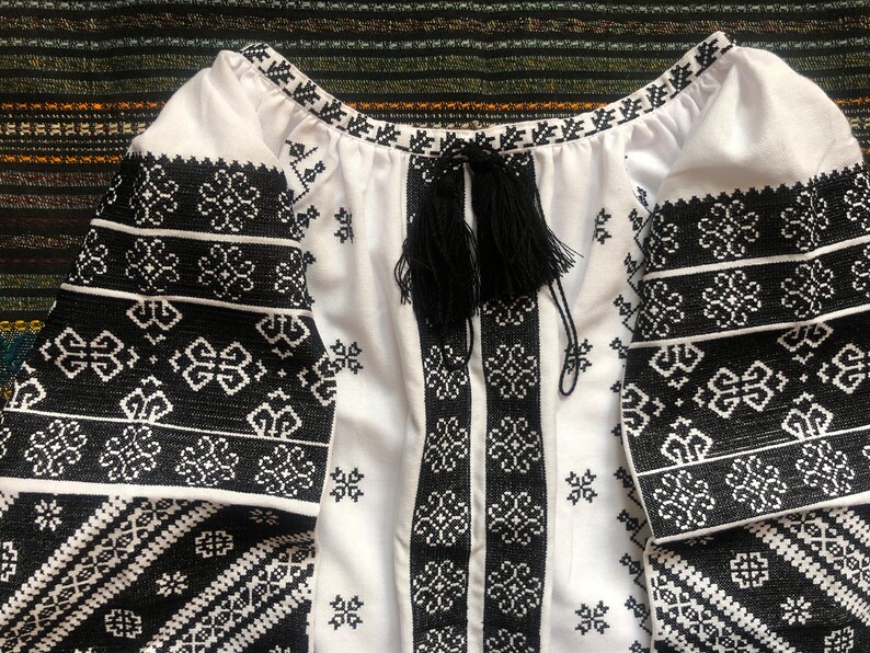 May include: White blouse with black embroidered floral patterns on the sleeves and bodice. The blouse features a round neckline and a black tassel tie. The sleeves are long and the fabric appears to be cotton or linen.
