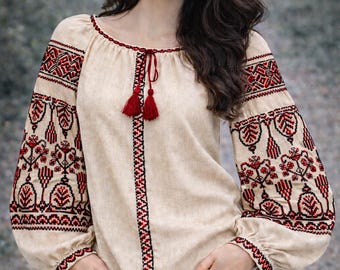 May include: Beige embroidered blouse with red and black floral patterns on the sleeves and bodice. The blouse features a round neckline with red tassels and long sleeves. A traditional folk garment.