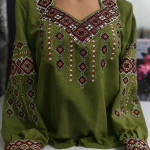 May include: Olive green long-sleeved blouse with detailed embroidery. The embroidery incorporates geometric patterns in red, white, and black, decorating the neckline, sleeves, and cuffs. The blouse has a V-neck and a relaxed fit, displaying a traditional design.