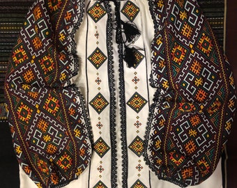 Puede incluir: Blusa blanca bordada con mangas largas y un patrón geométrico en tonos naranja, amarillo, verde y negro. La blusa tiene un ribete de encaje negro alrededor del cuello y las mangas.