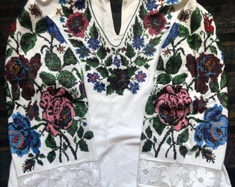 Puede incluir: Una blusa blanca bordada con un patrón floral en tonos de rosa, azul, verde y negro. El bordado se realiza con cuentas y cubre toda la parte delantera y las mangas de la blusa.