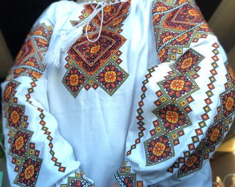 Puede incluir: Camisa blanca bordada con un patrón geométrico en naranja, rojo y verde. El bordado está en las mangas y en la parte delantera de la camisa.