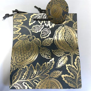 Puede incluir: Una bolsa de regalo negra con un estampado floral dorado y una etiqueta a juego. La bolsa tiene forma rectangular con un asa de cordón negro. La etiqueta es redonda y presenta un diseño floral dorado. El diseño es elegante.