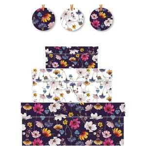 Puede incluir: Tres etiquetas de regalo redondas y tres cajas de regalo rectangulares con estampados florales. Las etiquetas están sujetas con hilo naranja. Las cajas están apiladas, cada una con un diseño floral diferente en tonos blancos, amarillos, rosas y azules sobre un fondo oscuro.