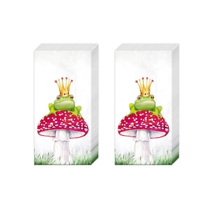 May include: Two stacks of white paper napkins. Each napkin features a green frog wearing a gold crown, sitting atop a red mushroom with white spots. The background shows green grass.