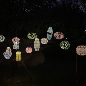 Puede incluir: Una colecci&oacute;n de faroles de papel coloridos y con intrincados patrones que cuelgan de las ramas de los &aacute;rboles en un entorno oscuro. Los faroles son de diversas formas y tama&ntilde;os, incluyendo redondos, ovalados y rectangulares.