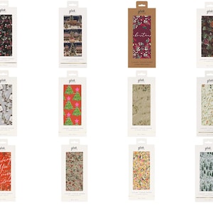 May include: A collection of twelve rectangular packages of Glick luxury tissue paper, each featuring distinct holiday-themed patterns including Christmas trees, festive foliage, winter scenes, and seasonal text, all displayed in individual white or brown card sleeves.
