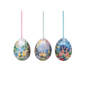 Puede incluir: Tres adornos de huevos de Pascua. Cada huevo presenta una escena pastoral diferente con un fondo de color pastel. Un huevo tiene patitos, otro conejitos y el tercero un diseño floral. Cada huevo está suspendido por una cinta fina.
