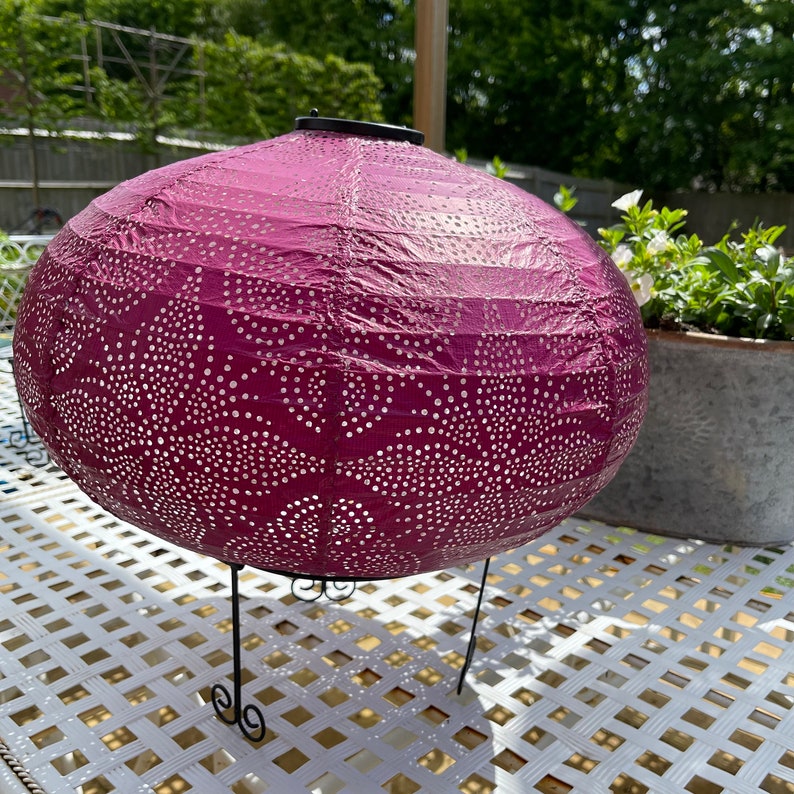 Puede incluir: Una linterna de papel morada con un patr&oacute;n de puntos blancos y un soporte de metal negro. La linterna est&aacute; iluminada por una luz solar.