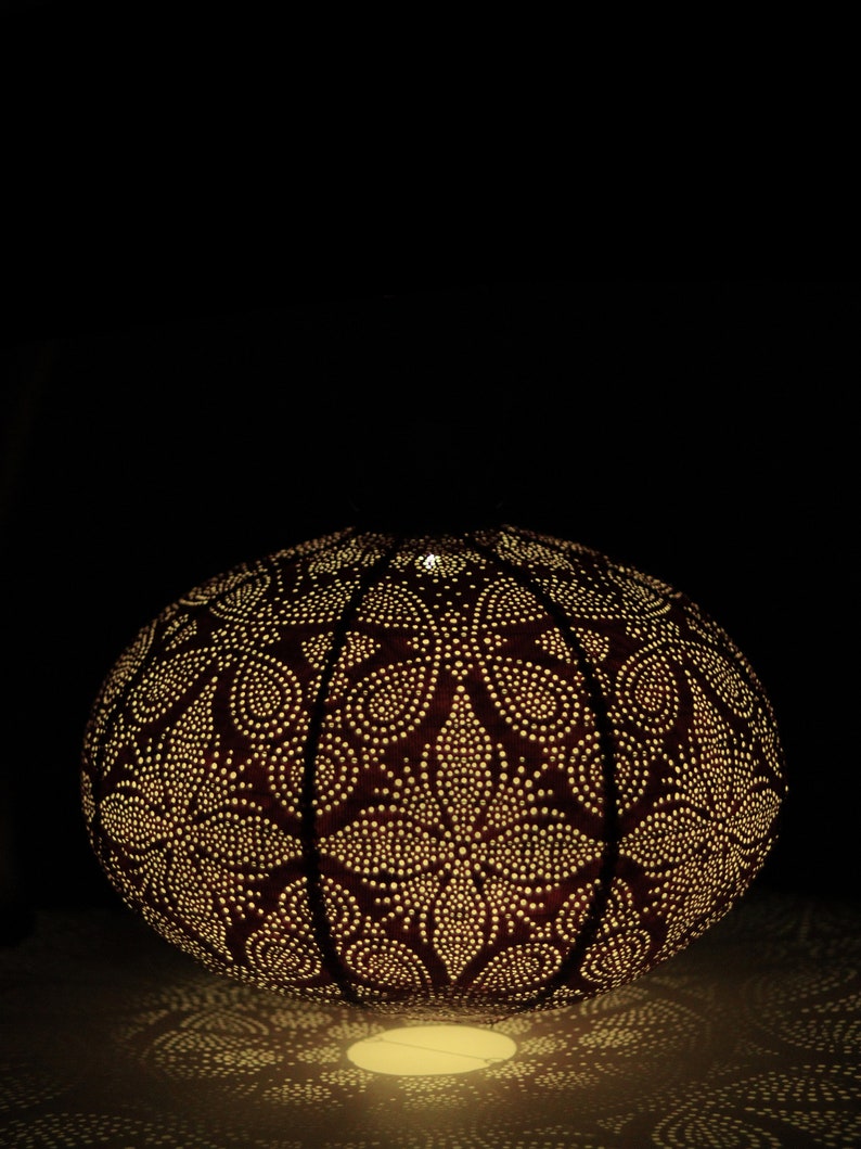 Puede incluir: Una pantalla de l&aacute;mpara redonda de metal marr&oacute;n oscuro con un intrincado patr&oacute;n floral. La pantalla est&aacute; iluminada desde el interior, proyectando un c&aacute;lido resplandor.