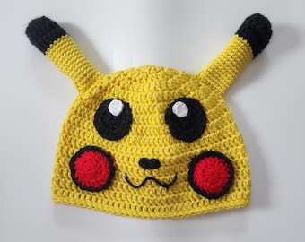 May include: Yellow crocheted hat with black ears and a Pikachu face. The hat has black eyes with white pupils, a black nose, and red cheeks.