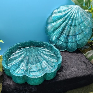 May include: A two-piece teal resin shell-shaped trinket dish with a lid. The surface of both pieces is translucent and embedded with fine glitter, creating a shimmering effect. The design features radial ridges mimicking a natural scallop shell.
