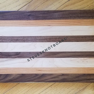 Puede incluir: Tabla de cortar rectangular de madera con franjas alternas de madera clara y oscura. La tabla presenta el texto "STEVESKNOTBOARD". Los tonos de madera incluyen beige claro, marrón medio y marrón oscuro.