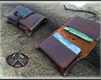 May include: Two brown leather pouches with white stitching. The larger pouch has a flap that opens to reveal a brown interior and a small pocket. The smaller pouch is closed and has a leather strap with a metal logo that reads "Indy Hammered Knives".