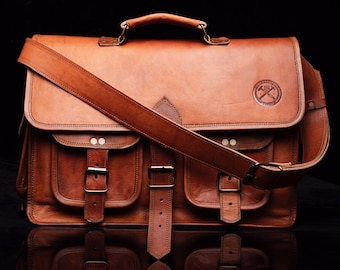 Op de afbeelding: Een bruine leren schoudertas met een grote klep en meerdere vakken. De tas heeft een lange verstelbare band en een metalen gesp. De tas heeft een logo aan de voorkant met de tekst "The Working Man's Supply Co."