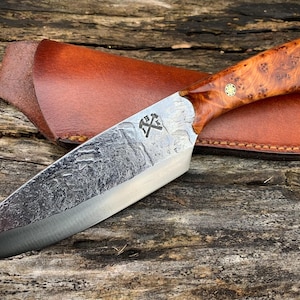 Könnte beinhalten: Ein handgemachtes Messer mit einem braunen Holzgriff und einer strukturierten Klinge aus Stahl. Das Messer befindet sich in einer braunen Lederscheide.