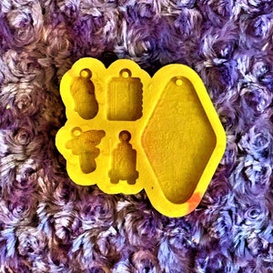 Könnte beinhalten: Gelbe Silikonform mit fünf verschiedenen Formen zum Herstellen von Schmuck. Die Formen umfassen ein Schild, eine Glocke, ein kleines Rechteck, einen kleinen Kreis und ein größeres Rechteck.