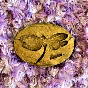 May include: A gold-coloured oval-shaped stone with a dragonfly design carved into the surface. The stone is sitting on a purple and white fuzzy surface.